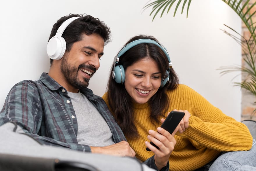 A couple sharing headphones, listening to a song together on a phone and smiling